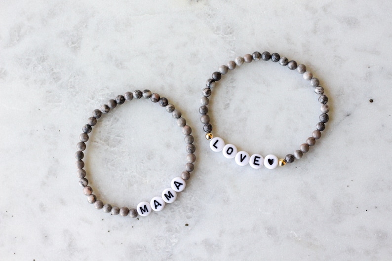 May include: Two beaded bracelets made of small, round, grey and white marbled stone beads. One bracelet features white letter beads spelling MAMA, while the other features white letter beads spelling LOVE followed by a small black heart symbol.