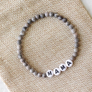 May include: A circular beaded bracelet featuring small, round, grey and brown mottled stone beads. The bracelet includes four white, flat, circular beads with the black printed letters M, A, M, and A, spelling out the word MAMA.