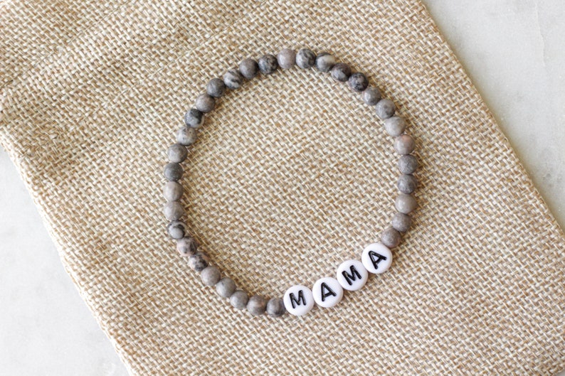 May include: A circular beaded bracelet featuring small, round, grey and brown mottled stone beads. The bracelet includes four white, flat, circular beads with the black printed letters M, A, M, and A, spelling out the word MAMA.