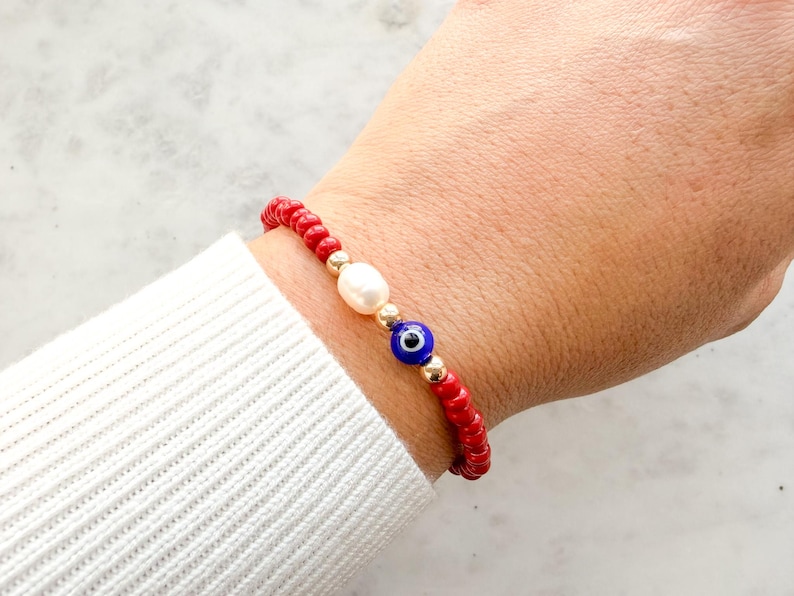 Peut inclure: Un bracelet de perles compos&eacute; de petites perles rouges sph&eacute;riques, d'une perle blanche ovale, d'une perle bleue et blanche en forme d'&oelig;il protecteur et de petites perles intercalaires m&eacute;talliques dor&eacute;es dispos&eacute;es en cercle.