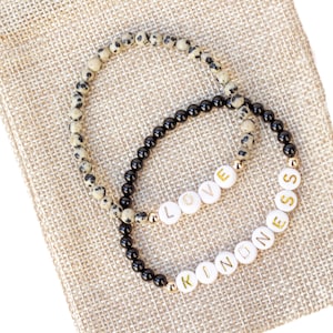 May include: Two beaded bracelets on a beige burlap background. One bracelet is black with black beads and the other is beige with black and white speckled beads. The bracelets spell out the words "LOVE" and "KINDNESS" in gold letters.