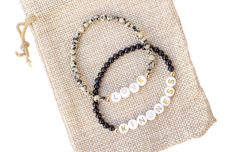 May include: Two beaded bracelets on a beige burlap background. One bracelet is black with black beads and the other is beige with black and white speckled beads. The bracelets spell out the words "LOVE" and "KINDNESS" in gold letters.