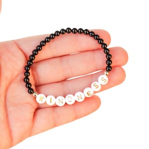 May include: Black beaded bracelet with white beads spelling out the word "KINDNESS" in gold lettering.