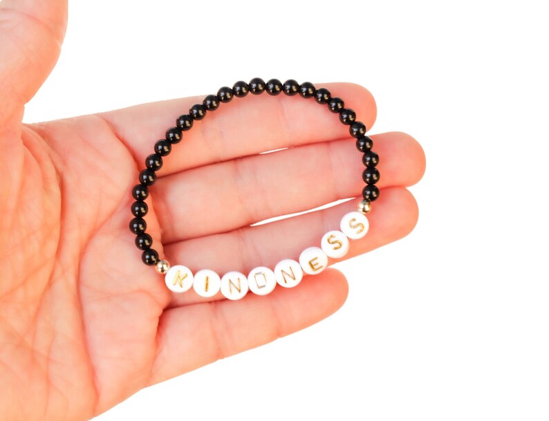May include: Black beaded bracelet with white beads spelling out the word "KINDNESS" in gold lettering.