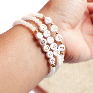May include: Three stacked beaded bracelets featuring pale pink translucent beads, small gold-toned metallic spacer beads, and white circular beads with gold-colored letters spelling out MAGGIE, MAMA, and J K, along with a small gold heart symbol.