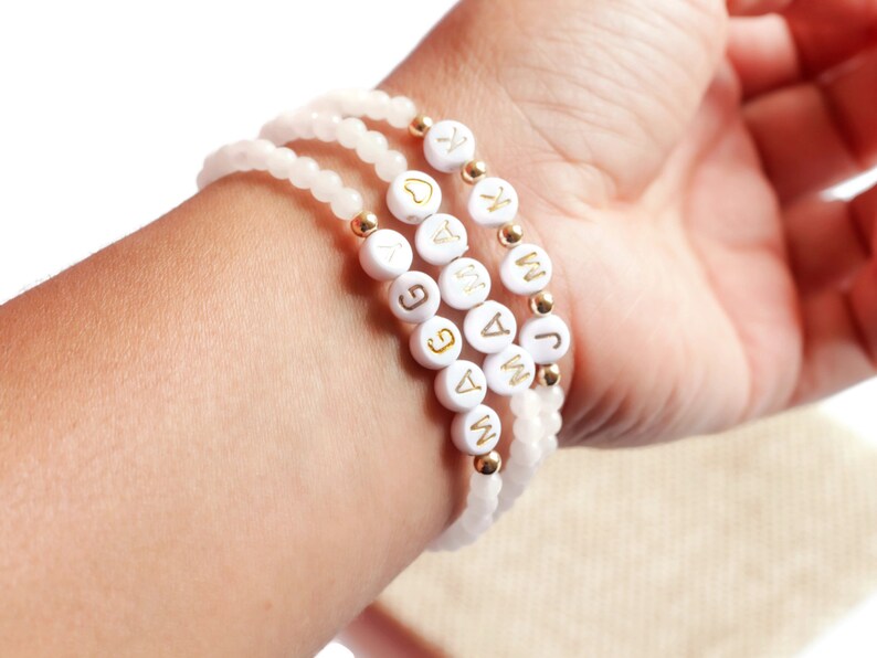 May include: Three stacked beaded bracelets featuring pale pink translucent beads, small gold-toned metallic spacer beads, and white circular beads with gold-colored letters spelling out MAGGIE, MAMA, and J K, along with a small gold heart symbol.