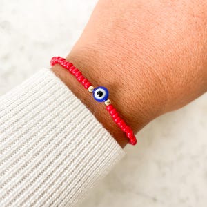 May include: Red beaded bracelet with a blue and white evil eye charm. The bracelet is made of small red beads and features a gold-colored accent bead on either side of the charm. The bracelet is worn on a wrist.