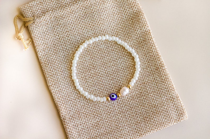 Peut inclure: Un bracelet de perles compos&eacute; de petites perles blanches translucides, d'une perle bleue et blanche en forme d'&oelig;il, d'une petite perle d'eau douce blanche et de deux minuscules perles intercalaires m&eacute;talliques dor&eacute;es.