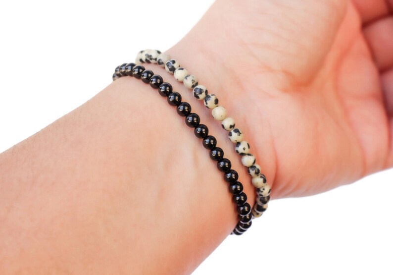 May include: Two bracelets on a wrist. One bracelet is made of black beads, the other is made of white and black speckled beads.