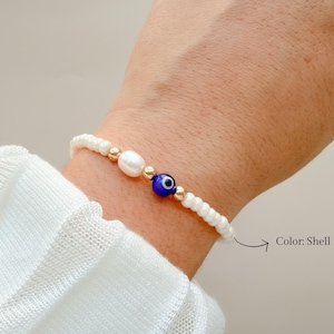 Peut inclure: Un bracelet de perles compos&eacute; de petites perles de coquillage blanches, d'une perle blanche ovale unique et d'une perle centrale bleue en forme d'&oelig;il. Le design inclut de petites perles intercalaires dor&eacute;es entre les &eacute;l&eacute;ments blancs et bleus. Le texte Color: Shell White est imprim&eacute; pr&egrave;s du bracelet.