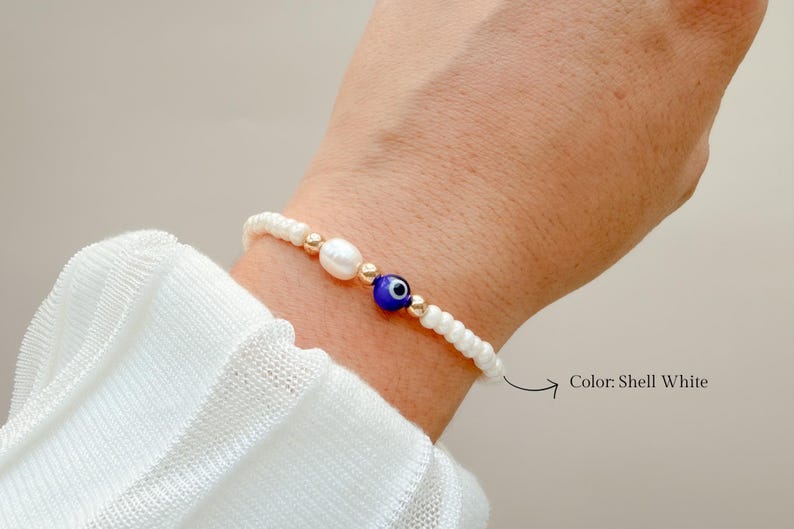 Peut inclure: Un bracelet de perles compos&eacute; de petites perles de coquillage blanches, d'une perle blanche ovale unique et d'une perle centrale bleue en forme d'&oelig;il. Le design inclut de petites perles intercalaires dor&eacute;es entre les &eacute;l&eacute;ments blancs et bleus. Le texte Color: Shell White est imprim&eacute; pr&egrave;s du bracelet.