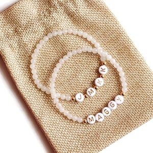 May include: Two white beaded bracelets with gold lettering spelling out the names "MAGGY" and "JMKY" in a burlap drawstring bag.