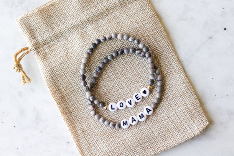 May include: Two stretch bracelets made of small, round, grey-toned mottled stone beads. One bracelet features white circular beads with black letters spelling LOVE and a black heart symbol. The other bracelet features white beads spelling MAMA.