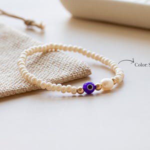 Peut inclure: Un bracelet de perles compos&eacute; de petites perles blanc coquillage, d'une perle &oelig;il bleu et blanc, d'une petite perle blanche et de perles intercalaires m&eacute;talliques dor&eacute;es. Le texte Color: Shell White est imprim&eacute; &agrave; c&ocirc;t&eacute; avec une fl&egrave;che.