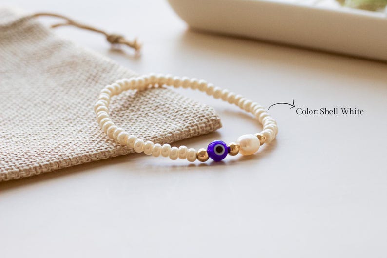 Peut inclure: Un bracelet de perles compos&eacute; de petites perles blanc coquillage, d'une perle &oelig;il bleu et blanc, d'une petite perle blanche et de perles intercalaires m&eacute;talliques dor&eacute;es. Le texte Color: Shell White est imprim&eacute; &agrave; c&ocirc;t&eacute; avec une fl&egrave;che.