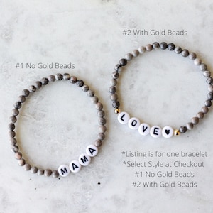 May include: Two beaded bracelets made of grey and brown mottled stone beads. One bracelet features white letter beads spelling MAMA. The other bracelet features white letter beads spelling LOVE with a heart symbol and small gold-colored accent beads.
