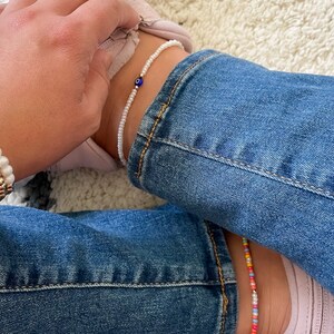 May include: A person wearing a stack of gold and white beaded bracelets with the letters "MAN" on them. The person is also wearing a white beaded anklet with a blue evil eye charm.