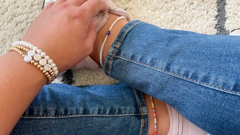 May include: A person wearing a stack of gold and white beaded bracelets with the letters "MAN" on them. The person is also wearing a white beaded anklet with a blue evil eye charm.