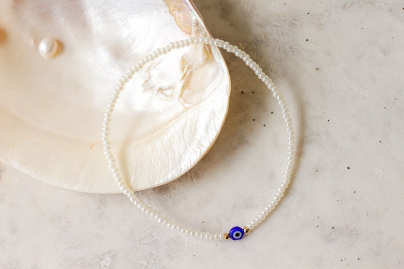 May include: A white pearl beaded bracelet with a blue evil eye charm. The bracelet is on a white background.