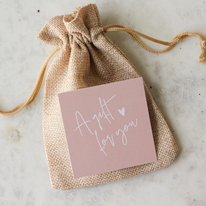 May include: A tan burlap drawstring pouch featuring a square dusty pink card attached to the front. The card displays the white handwritten text A gift for you with a small heart icon positioned above the word gift, centered on the pouch.