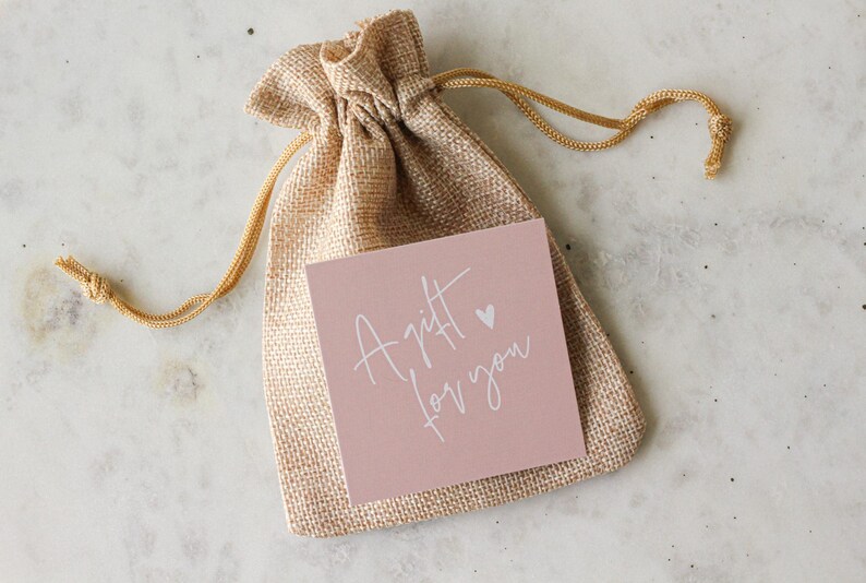 May include: A tan burlap drawstring pouch featuring a square dusty pink card attached to the front. The card displays the white handwritten text A gift for you with a small heart icon positioned above the word gift, centered on the pouch.