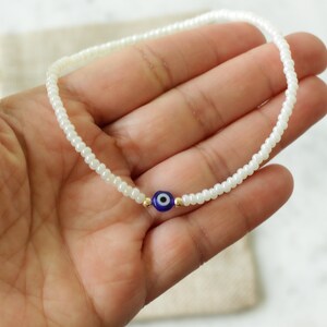 May include: White beaded bracelet with a blue evil eye charm. The bracelet is made of small white beads and has a single blue evil eye charm in the center.