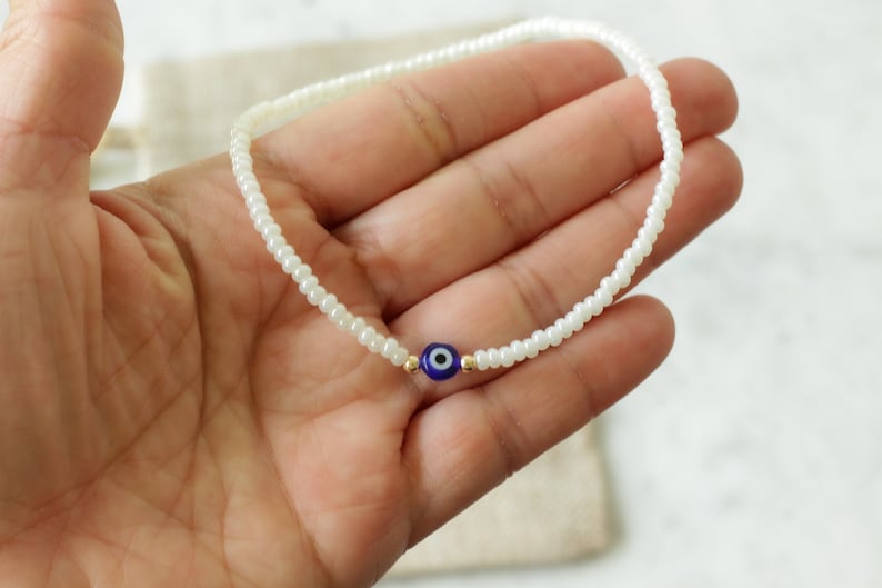 May include: White beaded bracelet with a blue evil eye charm. The bracelet is made of small white beads and has a single blue evil eye charm in the center.