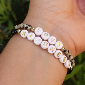 May include: Two bracelets with black and white beads spelling out the words "LOVE" and "KINDNESS" in gold letters.