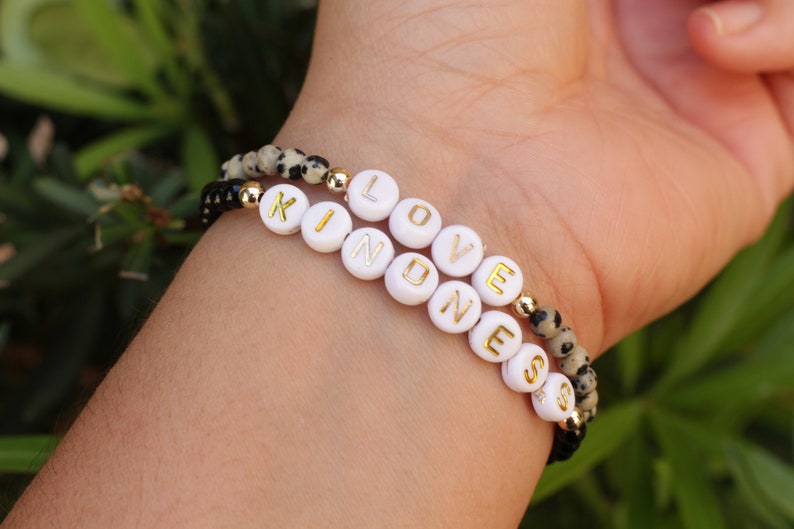 May include: Two bracelets with black and white beads spelling out the words "LOVE" and "KINDNESS" in gold letters.