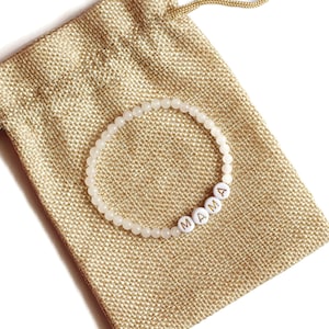 May include: A circular beaded bracelet featuring small, translucent, light-colored beads and four white spherical beads with the black printed letters M, A, M, and A, arranged to spell the word MAMA, resting on a tan, textured burlap drawstring pouch.