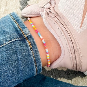 May include: A colorful beaded anklet with pink, yellow, blue, and white beads. The anklet is worn on a person's ankle above a pair of pink sneakers.