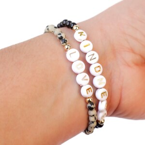 May include: Two bracelets on a wrist. One bracelet is black and white speckled and the other is white with gold lettering spelling out "LOVE" and "KINDNESS".