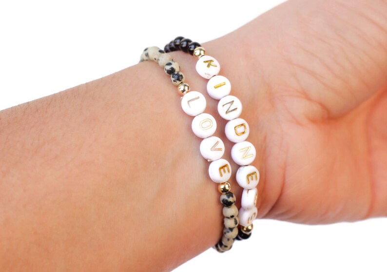May include: Two bracelets on a wrist. One bracelet is black and white speckled and the other is white with gold lettering spelling out "LOVE" and "KINDNESS".