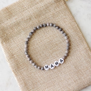 May include: A circular beaded bracelet featuring small, mottled grey and brown stone beads interspersed with four white, round, flat beads that spell out the word MAMA in black capital letters, resting on a tan, woven burlap drawstring pouch.