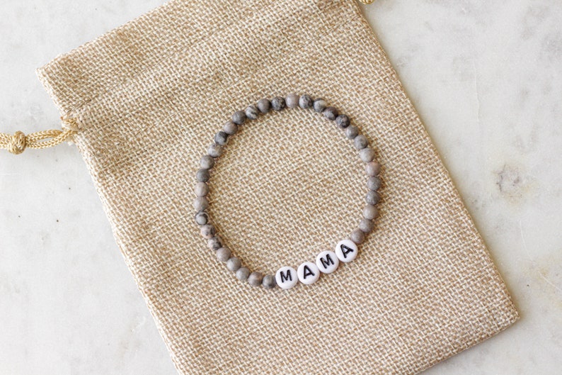 May include: A circular beaded bracelet featuring small, mottled grey and brown stone beads interspersed with four white, round, flat beads that spell out the word MAMA in black capital letters, resting on a tan, woven burlap drawstring pouch.