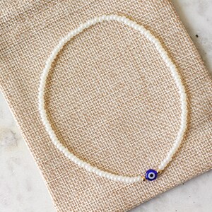 May include: A white beaded bracelet with a blue and gold evil eye charm. The bracelet is on a brown fabric background.