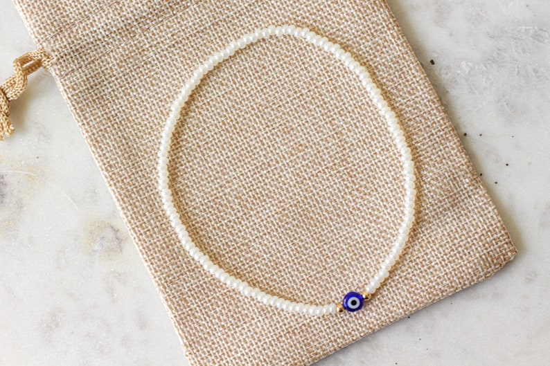 May include: A white beaded bracelet with a blue and gold evil eye charm. The bracelet is on a brown fabric background.