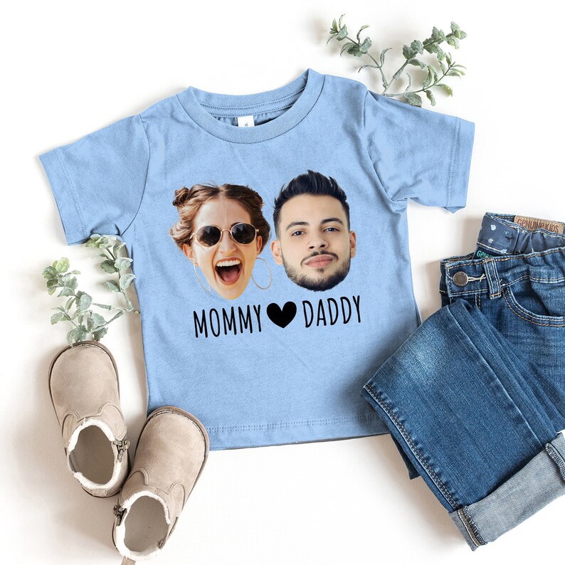 May include: A light blue t-shirt with a photo of a woman and a man's faces on it. The text "Mommy Daddy" is printed below the faces with a heart symbol between the words.