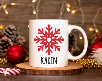 May include: White ceramic mug featuring a red snowflake design and the name "KAREN" in black lettering. The mug is set on a wooden coaster, with Christmas decorations in the background.
