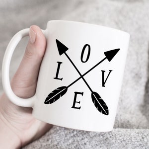 May include: White ceramic mug with a black design of two crossed arrows with feathers. The arrows form the word "LOVE" with the letters "O" and "V" in the centre of the design.