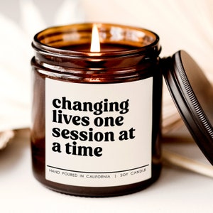 May include: A brown glass candle jar with a white label that reads "changing lives one session at a time". The candle is lit and the label also says "Hand Poured in California | Soy Candle".