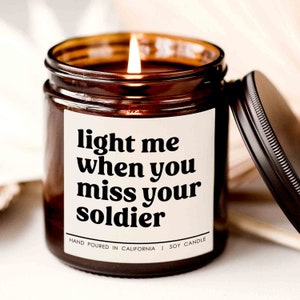 May include: A brown glass candle jar with a white label that reads "light me when you miss your soldier". The candle is lit and the label also says "Hand Poured in California | Soy Candle".