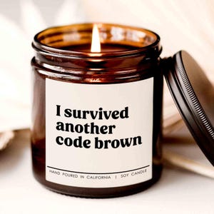 May include: A brown glass candle with a white label that reads "I survived another code brown". The candle is hand-poured in California and is a soy candle.