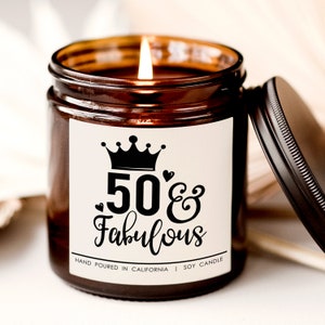 May include: A brown glass candle jar with a white label that says "50 & Fabulous" with a crown above the text. The candle is lit and the jar has a black lid.