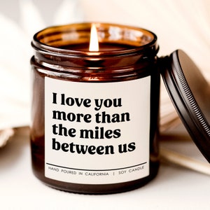 May include: A brown glass candle jar with a white label that reads "I love you more than the miles between us". The candle is lit and the label also says "Hand poured in California | Soy Candle".