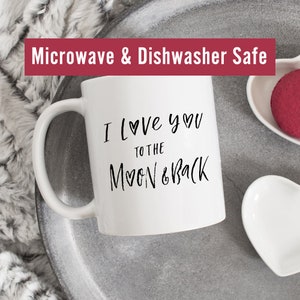 May include: White ceramic mug with black text that reads "I love you to the moon & back" with a heart symbol in place of the "o" in love and moon. The mug is on a gray tray with a white heart-shaped dish and a red heart-shaped object. The text "Microwave & Dishwasher Safe" is at the top of the image.