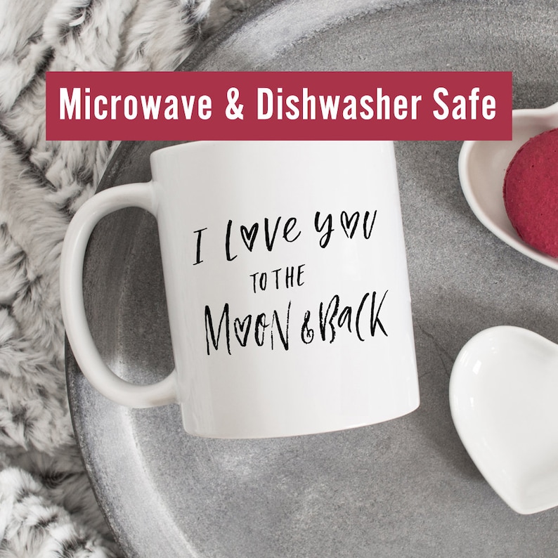 May include: White ceramic mug with black text that reads "I love you to the moon & back" with a heart symbol in place of the "o" in love and moon. The mug is on a gray tray with a white heart-shaped dish and a red heart-shaped object. The text "Microwave & Dishwasher Safe" is at the top of the image.