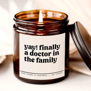 May include: A brown glass candle with a white label that reads "yay! finally a doctor in the family". The candle is hand-poured in California and made with soy wax.