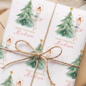 May include: Gift-wrapped present with white paper featuring a watercolor design of a ballerina, Christmas tree, and gold star. The text "Merry Christmas Madison" is printed in pink. The gift is tied with twine and rests on a brown paper package.