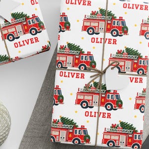 May include: White wrapping paper featuring a repeating pattern of red fire trucks carrying Christmas trees on their roofs, interspersed with the name Oliver in red text, tied with brown twine and accented by white star and heart gift tags.